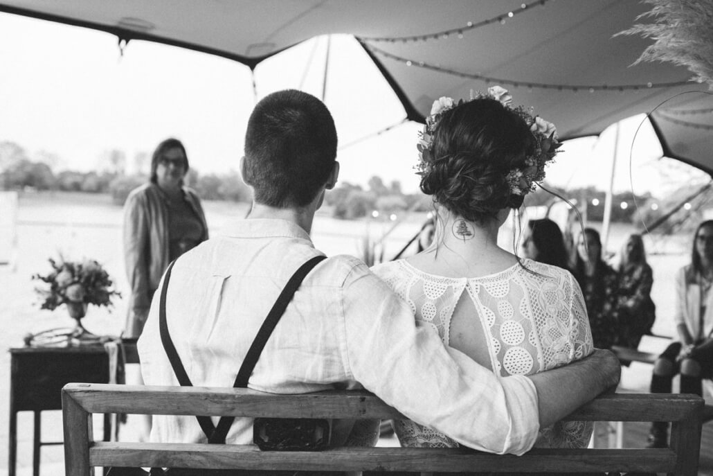 Brautpaar von hinten sitzend auf einer Holzbank mit Traurednerin unter einem Zelt